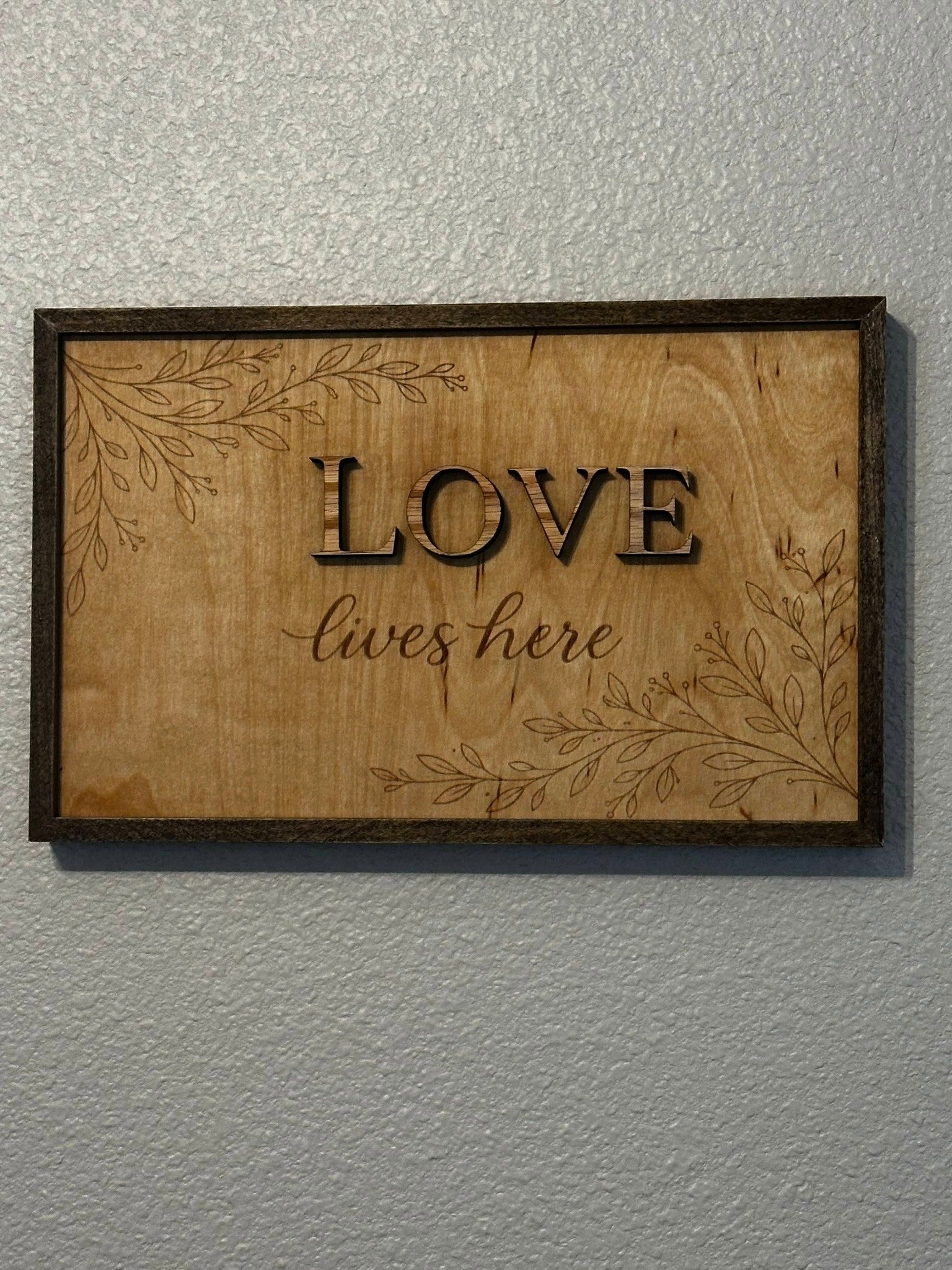 Love Lives Here framed wood sign with engraved botanical accents and dark frame hanging on wall