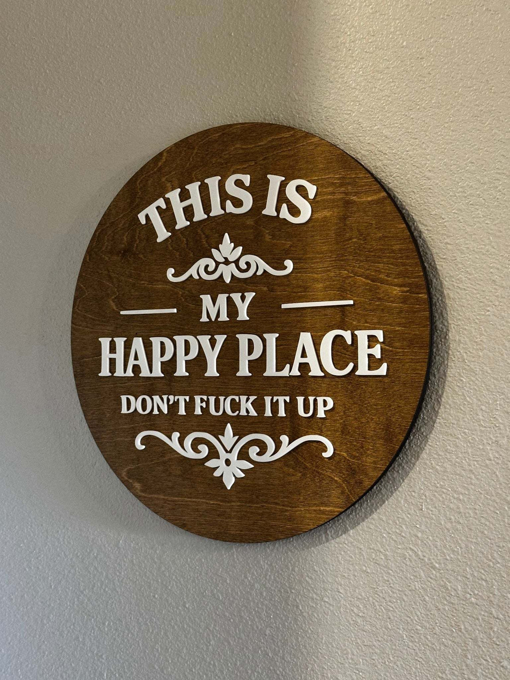 Round wood sign with raised white lettering reading "This Is My Happy Place — Don't F*ck It Up" on stained wood backer with decorative scroll accents