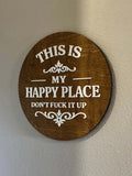 Round wood sign with raised white lettering reading "This Is My Happy Place — Don't F*ck It Up" on stained wood backer with decorative scroll accents
