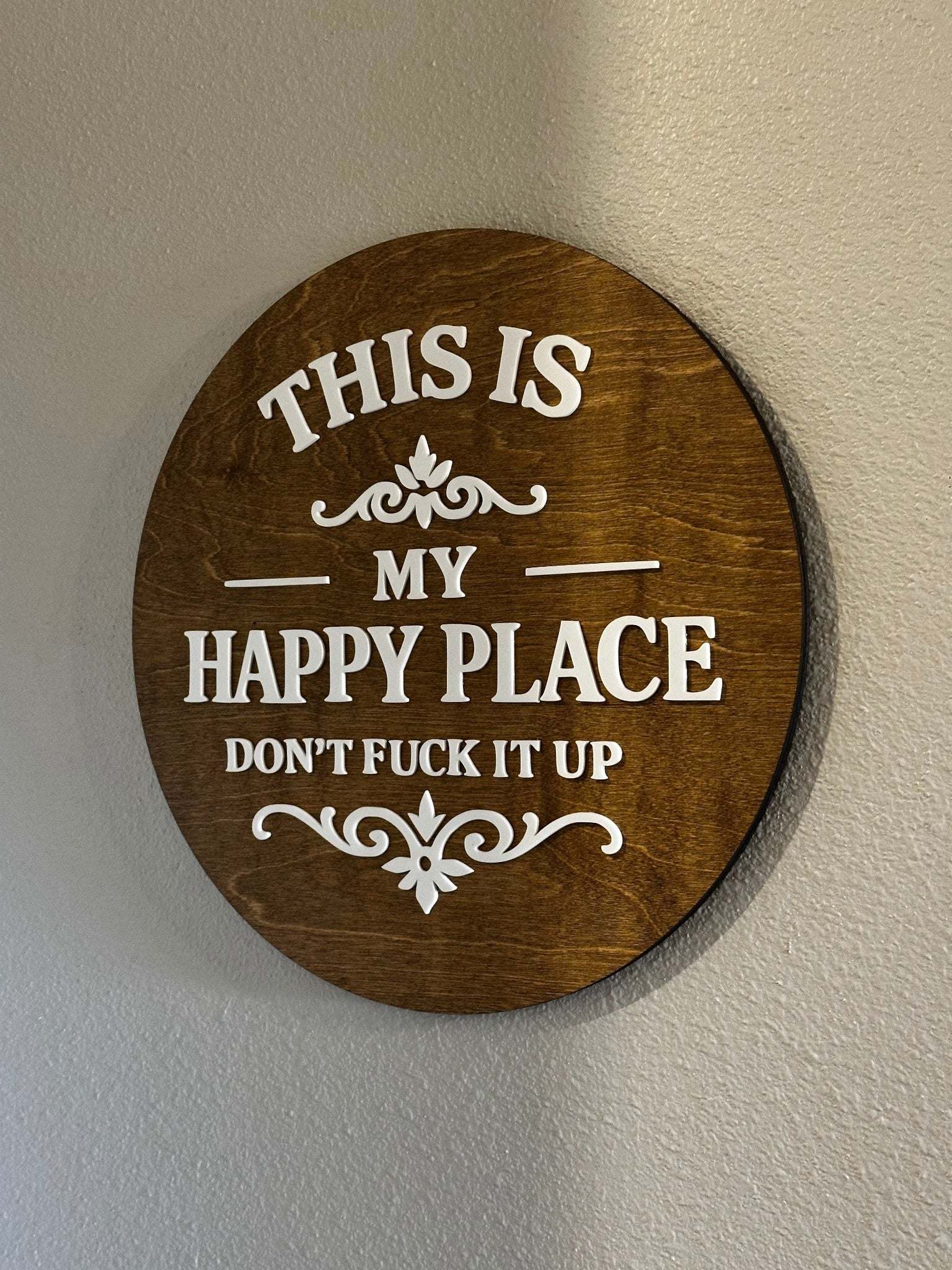 Round wood sign with raised white lettering reading "This Is My Happy Place — Don't F*ck It Up" on stained wood backer with decorative scroll accents