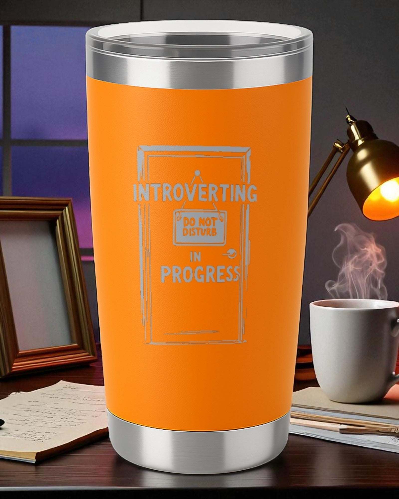 20oz orange engraved stainless steel tumbler with "Introverting in Progress" design and clear slider lid on desk with books and lamp