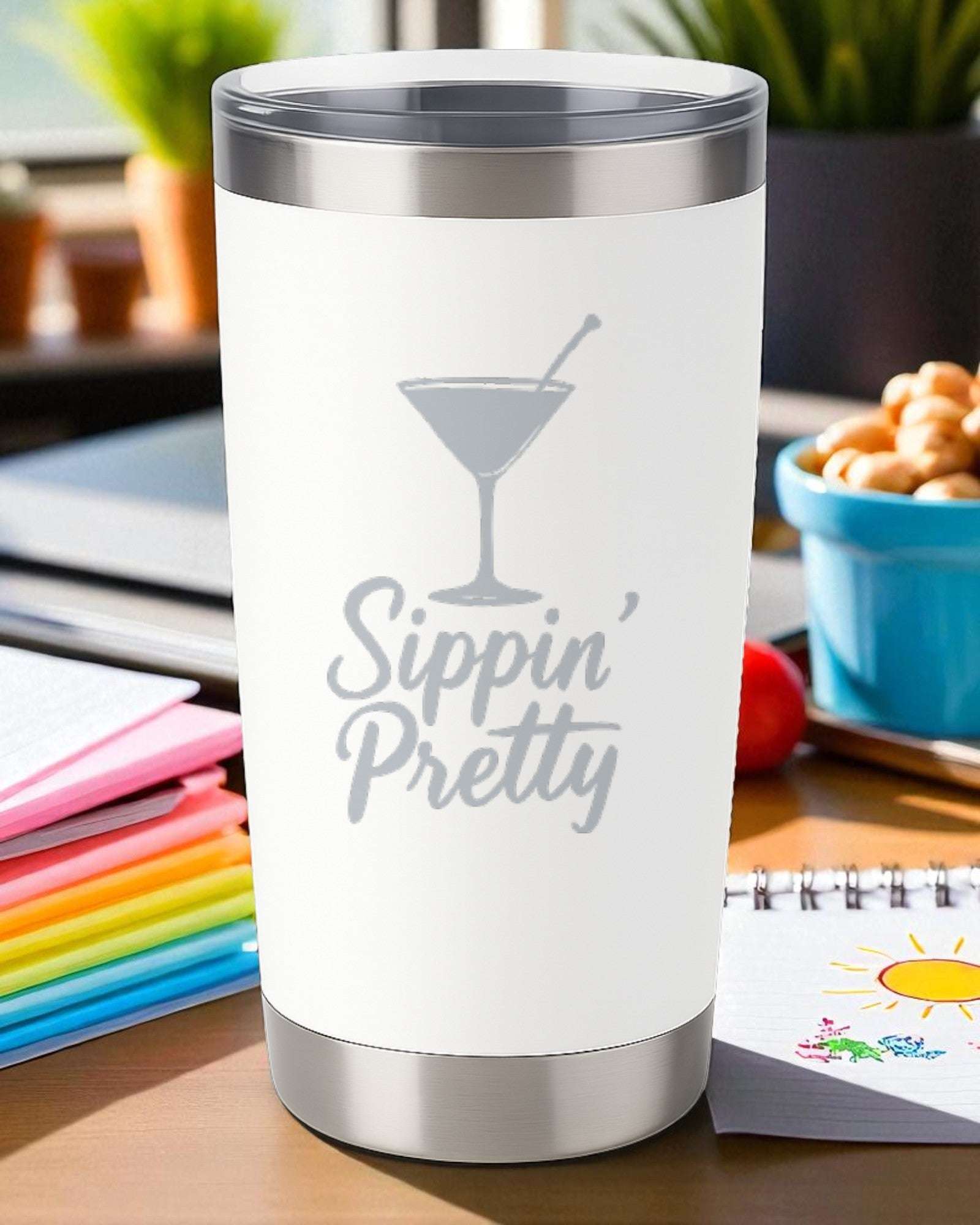 White 20oz stainless steel tumbler with "Sippin’ Pretty" engraved and clear lid on a table with colorful notebooks and snacks.